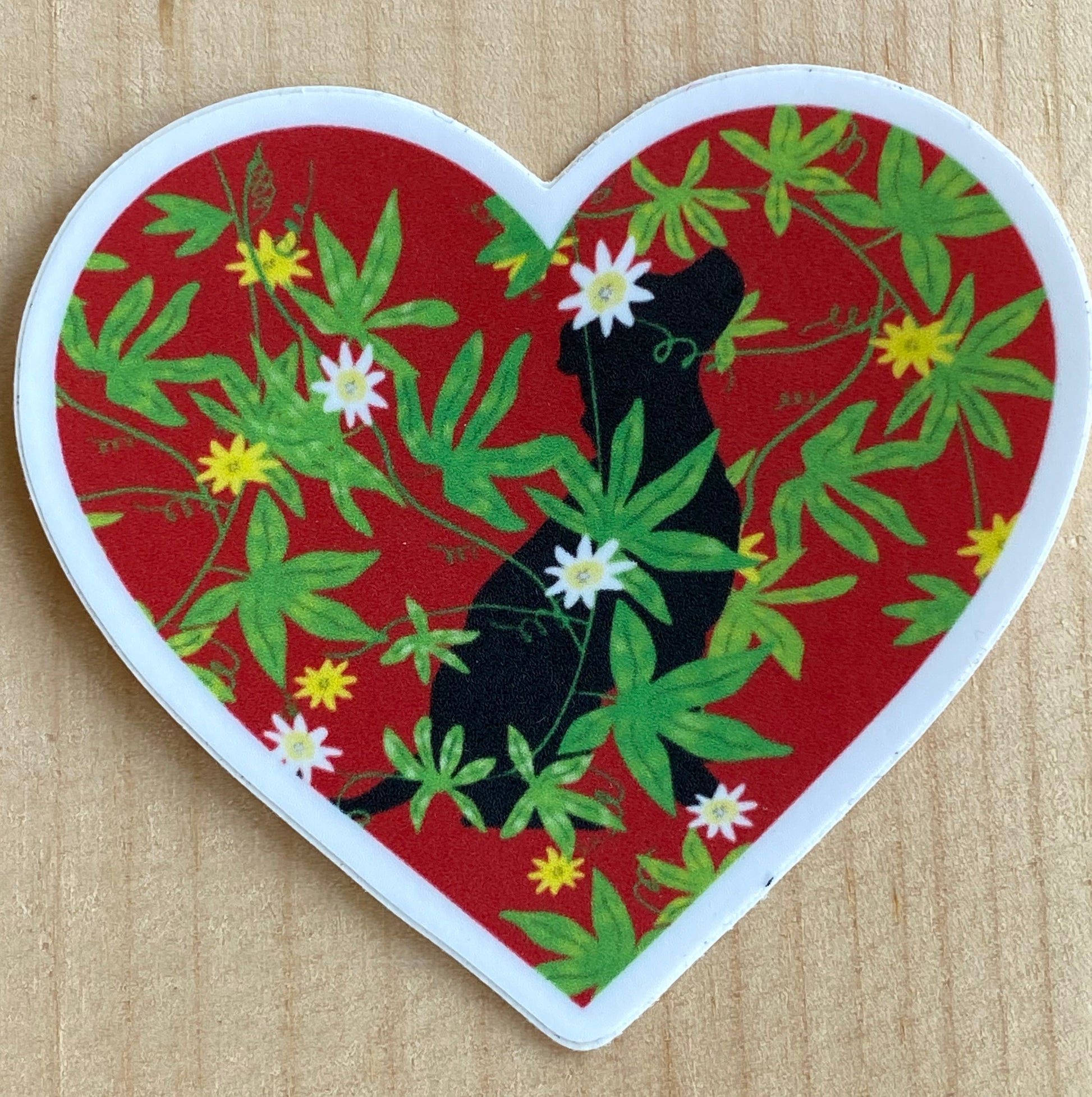 Heart-shaped sticker with black dog and green leaves on a red background, placed on a wooden surface.